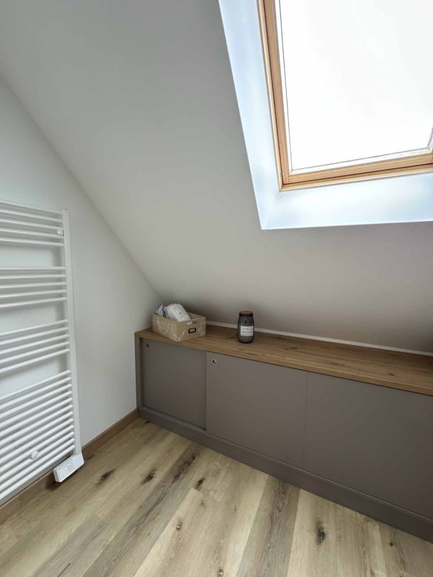 Meuble bas sur mesure en finition grise et bois clair, installé sous une fenêtre de toit, avec un radiateur mural à proximité et un sol en parquet chaleureux.