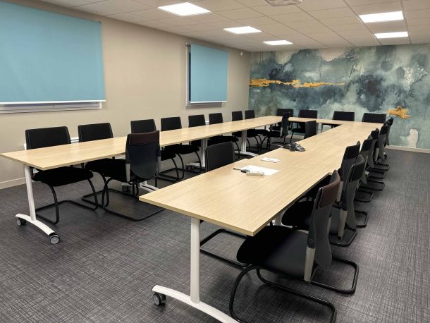 Salle de réunion spacieuse équipée de longues tables modulables en bois clair, entourées de chaises noires, avec un mur décoratif à motifs aquatiques bleus et dorés, et des stores bleus sur les fenêtres.
